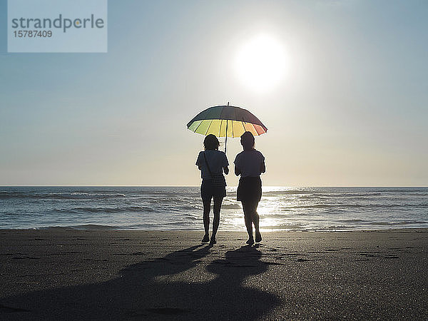 Zwei Frauen mit Sonnenschirm  die in der Abenddämmerung am Strand spazieren gehen  Kedungu-Strand  Bali  Indonesien