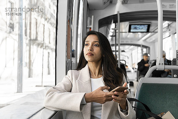 Junge Frau mit Smartphone  die in einer Straßenbahn aus dem Fenster schaut