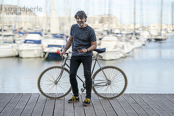 Porträt eines lächelnden  reifen Mannes mit Fixie-Fahrrad und Handy an der Strandpromenade  Alicante  Spanien