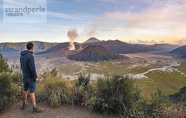 Junger Mann vor Vulkanlandschaft bei Sonnenuntergang  Blick in Tengger Caldera  rauchender Vulkan Gunung Bromo  im Vordergrund Mt. Batok  im Hintergrund Mt. Kursi  Mt. Gunung Semeru  Nationalpark Bromo-Tengger-Semeru  Java  Indonesien  Asien