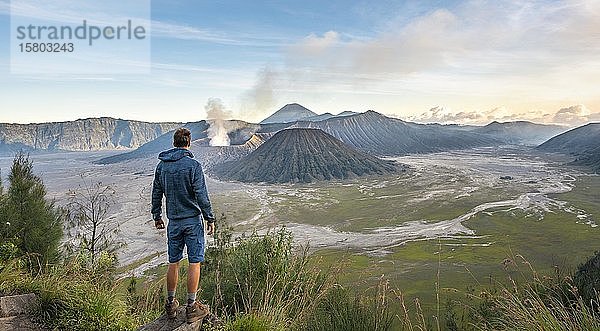 Junger Mann vor Vulkanlandschaft  Blick in Tengger Caldera  rauchender Vulkan Gunung Bromo  davor Mt. Batok  dahinter Mt. Kursi  Mt. Gunung Semeru  National Park Bromo-Tengger-Semeru  Java  Indonesien  Asien