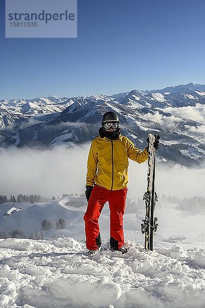 Skifahrer stehend an der Skipiste mit Ski in der Hand  Blick in die Kamera  verschneites Bergpanorama  Gipfel Hohe Salve  SkiWelt Wilder Kaiser Brixenthal  Hochbrixen  Tirol  Österreich  Europa