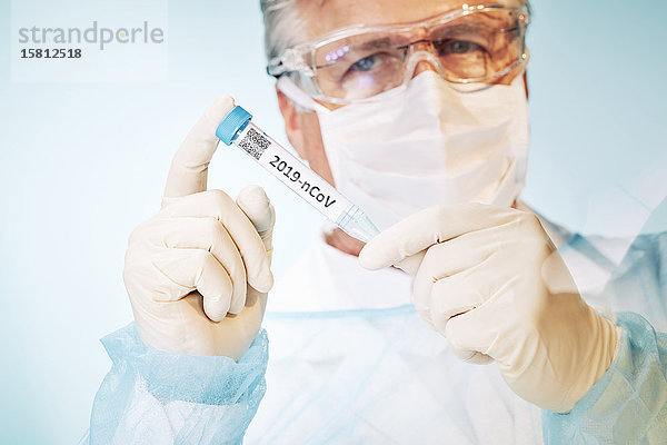 Grauhaariger Laborant mit Schutzbrille und Mundschutz blickt auf Reagenzglas mit Cornoravirus Probe