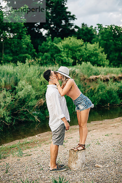 Junges Paar steht an einem Flussufer und küsst sich.