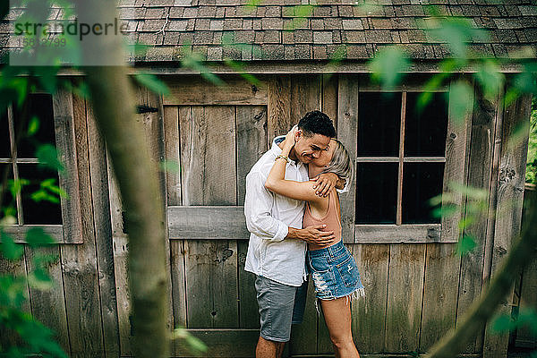Junges Paar steht vor einer Holzhütte  umarmt und küsst sich.