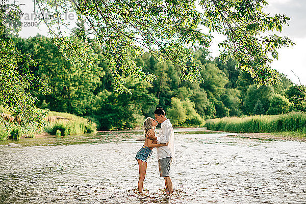 Junges Paar steht knöcheltief in einem Fluss  umarmt und küsst sich.