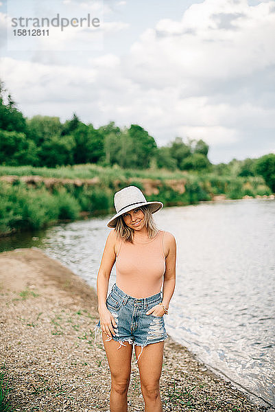 Junge blonde Frau in Shorts und mit Hut  die an einem Flussufer steht und in die Kamera lächelt.