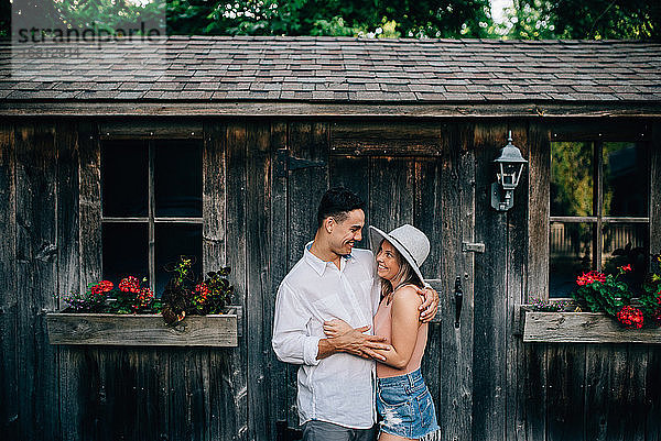 Junges Paar steht vor einer Holzhütte  umarmt sich und lächelt einander an.