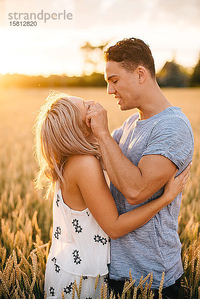 Lächelndes junges Paar steht im goldenen Weizenfeld und lächelt sich an.