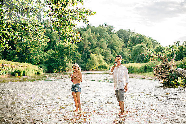 Junges Paar  das knöcheltief in einem Fluss steht und mit dem Handy fotografiert.