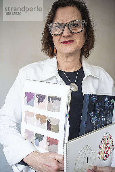 Bildnis einer brünetten Frau mit Brille und weißer Jeansjacke  die Skizzenbücher des Künstlers in der Hand hält.