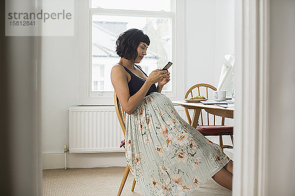 Schwangere Frau im geblümten Kleid benutzt ihr Smartphone am Tisch