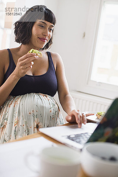 Schwangere Frau isst am Laptop