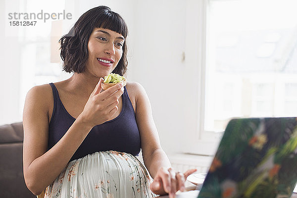 Glückliche schwangere Frau isst Avocado-Toast am Laptop