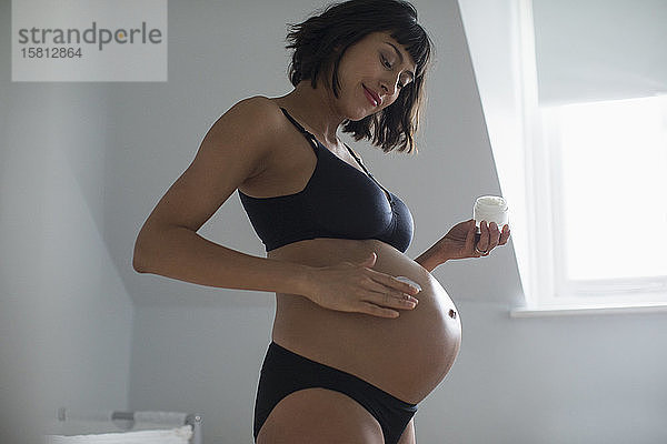 Schwangere Frau in BH und Höschen trägt Feuchtigkeitscreme auf den Bauch auf