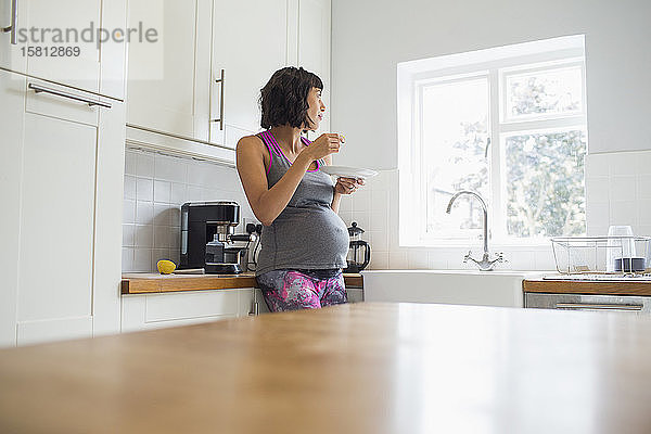 Nachdenkliche schwangere Frau  die in der Küche isst und aus dem Fenster schaut