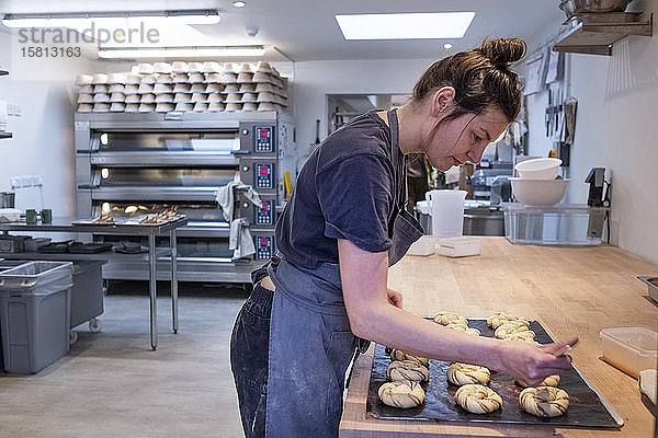 Frau mit Schürze  die in einer handwerklichen Bäckerei steht und Zimtbrötchen auf ein Backblech streicht.