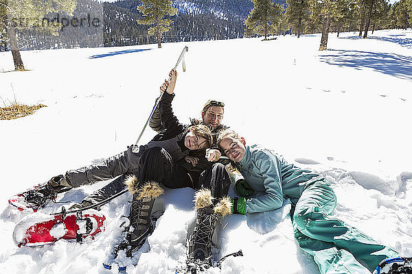 Eine Frau und ihre zwei Kinder  ein Mädchen im Teenageralter und ein Junge  die in Schneeschuhen und Skiausrüstung im Schnee liegen.