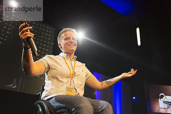 Lächelnde  selbstbewusste Rednerin mit Mikrofon im Rollstuhl auf der Bühne