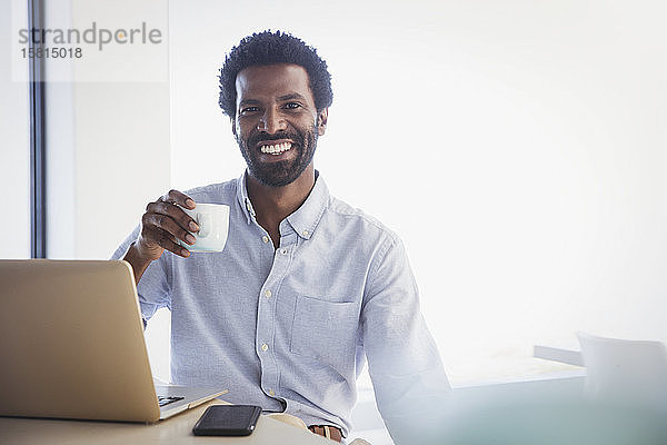 Porträt eines lächelnden  selbstbewussten Geschäftsmannes  der am Laptop Kaffee trinkt