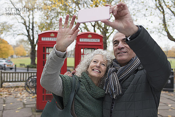 Lächelndes älteres Paar  das ein Selfie im Herbstpark vor einer roten Telefonzelle macht