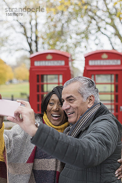 Glückliches älteres Paar  das ein Selfie vor einer roten Telefonzelle im Herbstpark macht