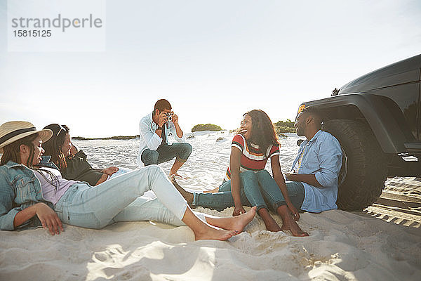 Junger Mann mit Digitalkamera  der seine Freunde am sonnigen Strand fotografiert