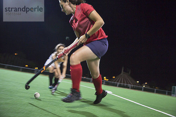 Junge Feldhockeyspielerinnen rennen um den Ball und spielen nachts auf dem Feld