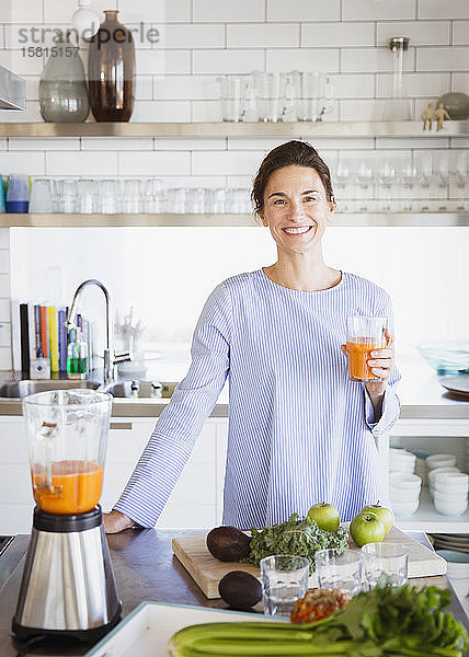 Porträt einer lächelnden  zuversichtlichen brünetten Frau  die gesunden Karottensaft an einem Mixer in der Küche trinkt