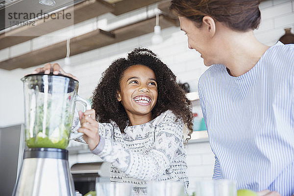 Enthusiastische Tochter hilft ihrer Mutter bei der Zubereitung eines gesunden grünen Smoothies im Mixer in der Küche