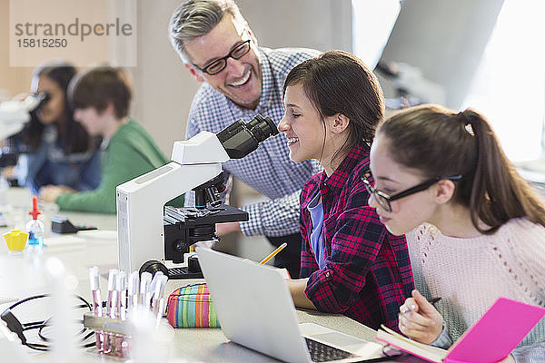 Lächelnder männlicher Lehrer der Naturwissenschaften hilft Schülerinnen bei der Durchführung eines wissenschaftlichen Experiments am Mikroskop im Labor