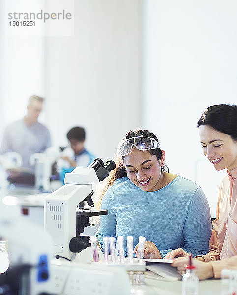 Lächelnde Lehrerin der Naturwissenschaften und Schülerin  die ein Lehrbuch am Mikroskop im Laboratorium liest