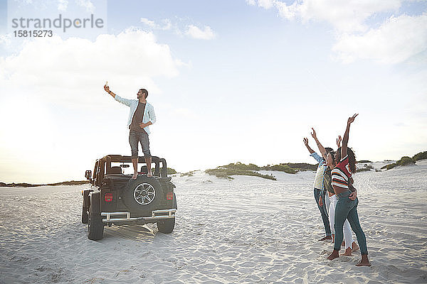Junger Mann auf Jeep-Reifen macht Selfie mit Freunden am Strand