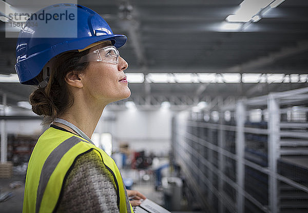 Lächelnde  zuversichtliche weibliche Aufsichtsperson mit Schutzhelm  die in einer Fabrik nach oben schaut