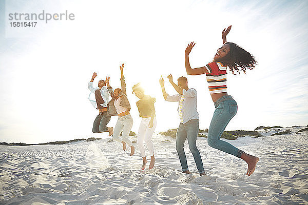 Portrait verspielte  energiegeladene junge Freunde  die am sonnigen Sommerstrand vor Freude springen