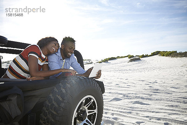 Junges Paar  das ein digitales Tablet im Jeep am sonnigen Strand benutzt und einen Roadtrip genießt