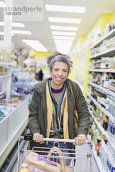 Porträt einer lächelnden  selbstbewussten Frau  die einen Einkaufswagen in einem Supermarktgang schiebt