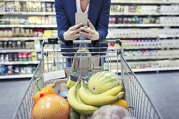 Frau mit Smartphone schiebt Einkaufswagen im Supermarkt