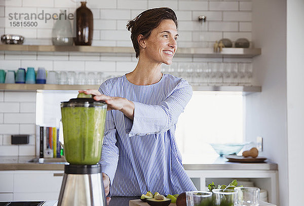 Lächelnde brünette Frau macht gesunden grünen Smoothie im Mixer in der Küche