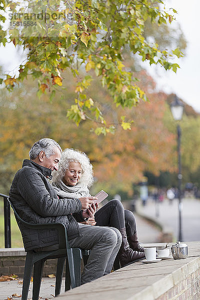 Älteres Paar  das ein digitales Tablet benutzt und Kaffee und Tee im Herbstpark genießt