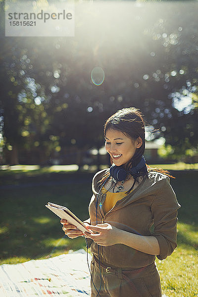 Lächelnde junge Frau mit Kopfhörern und digitalem Tablet in einem sonnigen Sommerpark