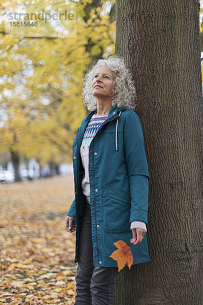 Gelassene ältere Frau lehnt an einem Baum im herbstlichen Park
