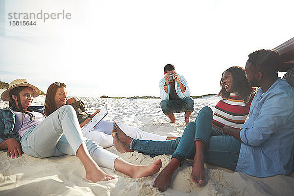 Junger Mann mit Digitalkamera fotografiert Freunde beim Entspannen am Strand