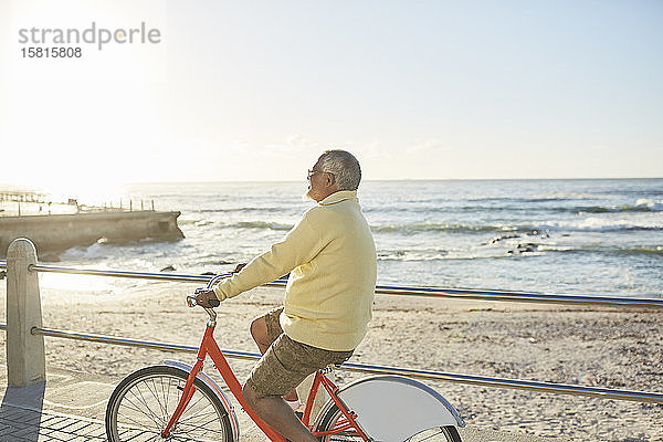 Aktiver älterer Mann  der auf der sonnigen Strandpromenade am Meer Fahrrad fährt