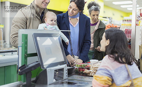 Freundliche Kassiererin im Gespräch mit und Hilfe für ein Paar mit Baby an der Supermarktkasse