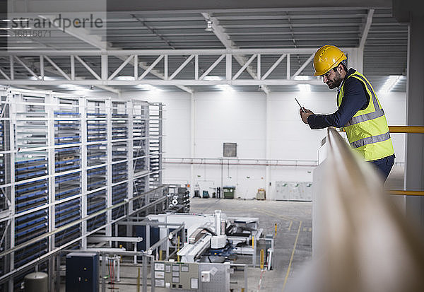 Männlicher Aufseher mit Klemmbrett auf einer Plattform  der auf eine Fabrik hinunterblickt