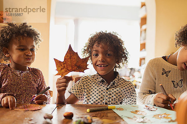 Portrait glücklicher Junge mit Herbstblatt beim Basteln mit Schwestern am Tisch