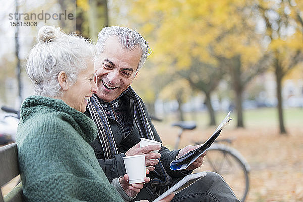 Lächelndes älteres Paar liest Zeitung und trinkt Kaffee auf einer Bank im Herbstpark