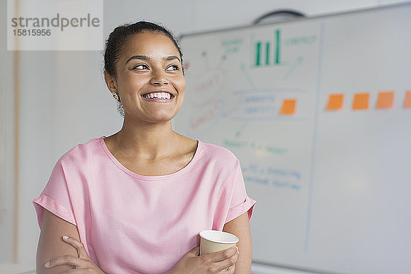 Porträt einer lächelnden  ehrgeizigen Geschäftsfrau  die am Whiteboard im Büro Kaffee trinkt