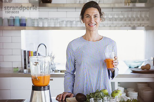 Porträt einer selbstbewussten  lächelnden Frau  die in der Küche gesunden Karottensaft trinkt